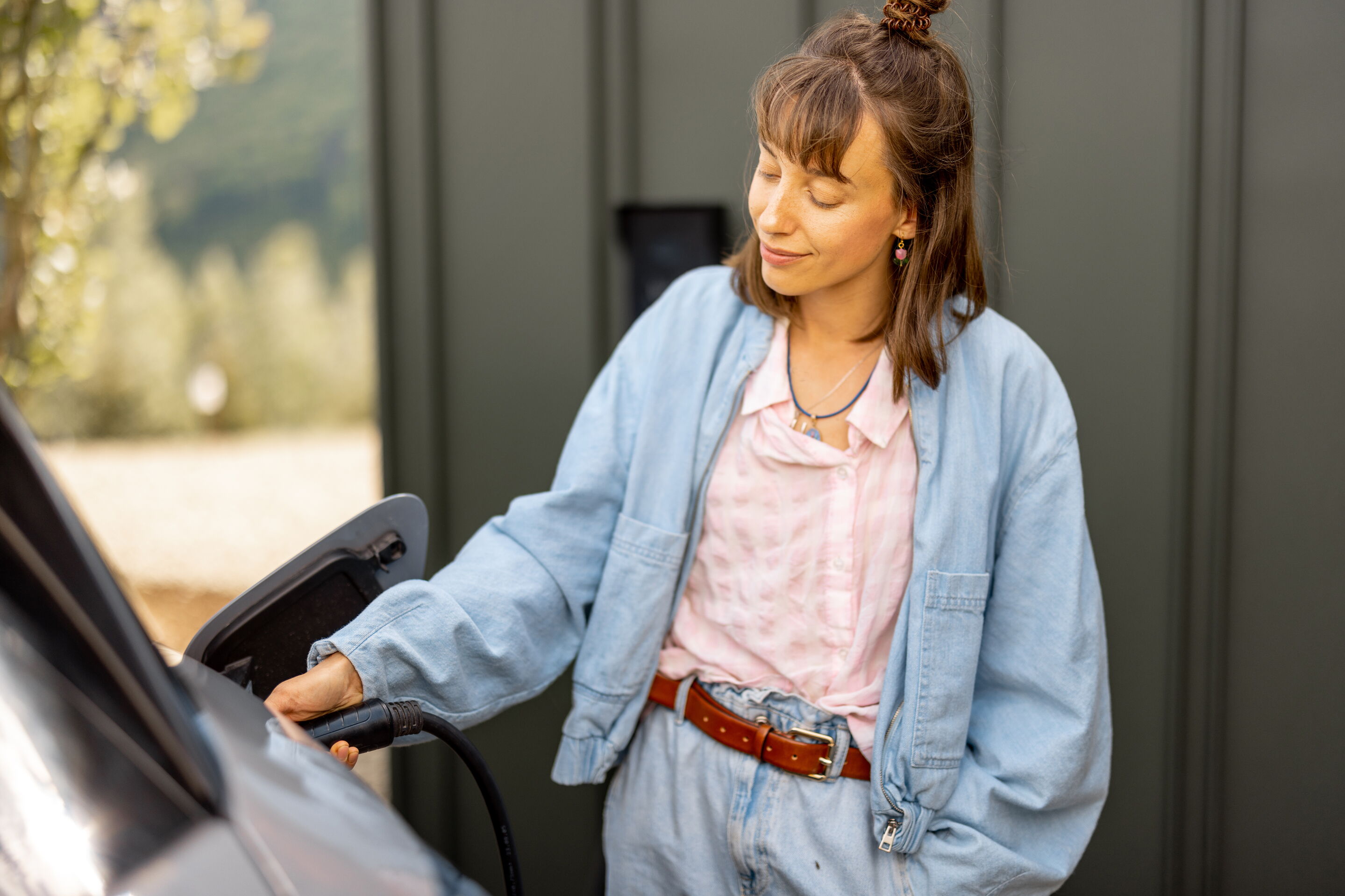 Das Motiv zeigt eine junge Frau in einer blauen Jeanskombi. Sie steht neben Ihrem E-mobil lädt Ihr Fahrzeug an einer Wallbox.