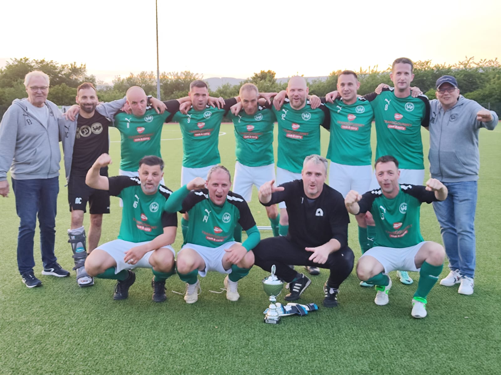 Auf dem Bild ist eine Fußballmannschaft der Spielvereinigung Ingelheim zu sehen. Die Spieler tragen ein grünes Trikot und stehen in zwei Reihen. Die Männer in der vorderen Reihe sind in der Hocke und strecken die Fäuste siegerbringend in die Luft.
