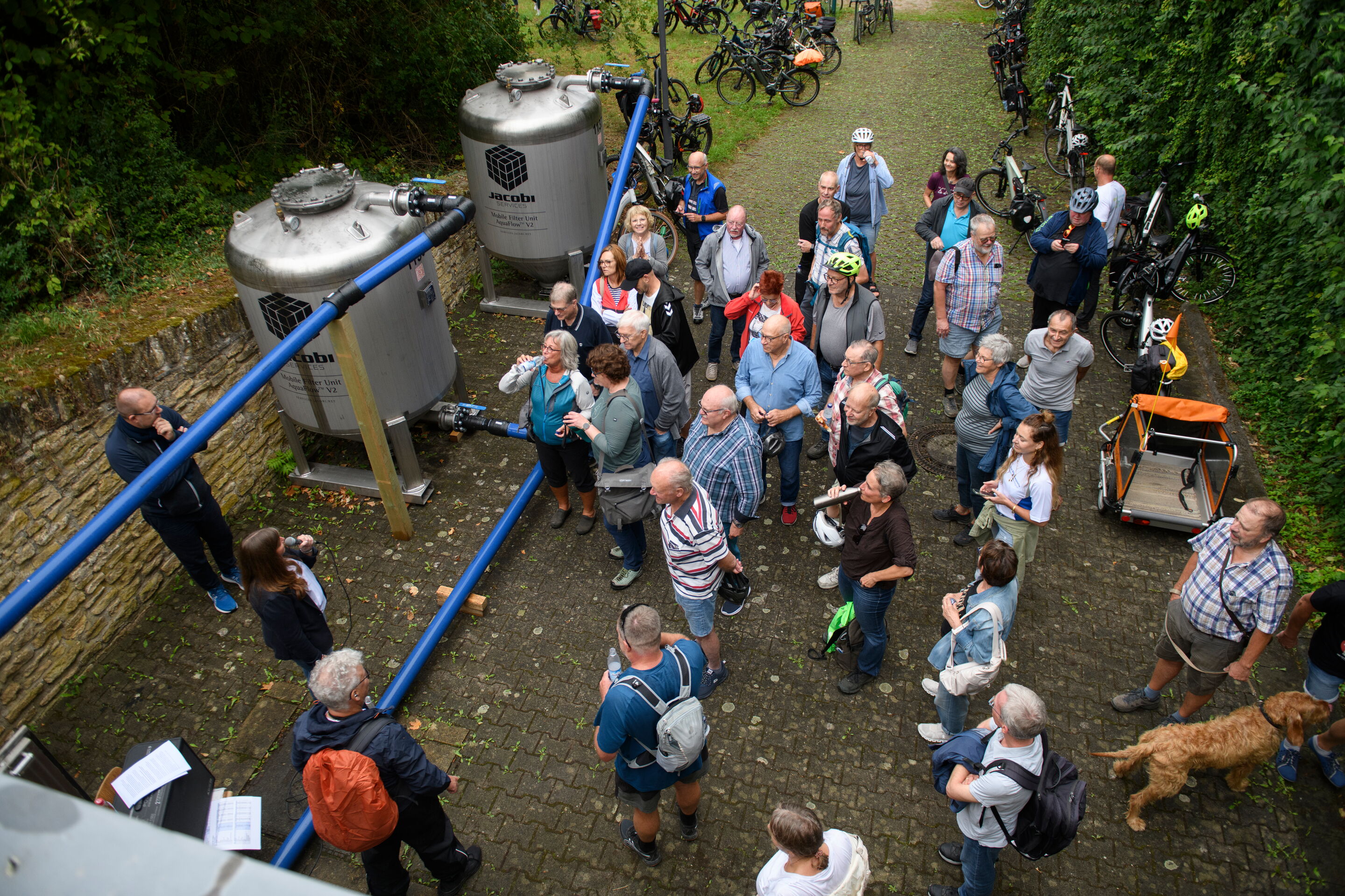 Das Bild zeigt zwei PFAS-Aktivkohlefilter in Heidesheim. Das Bild wurde beim Wasserradeln-Tag aufgenommen. Zu sehen sind außerdem etliche Zuhörer beim Vortrag von Lisa Gehrig zu den Filtern.