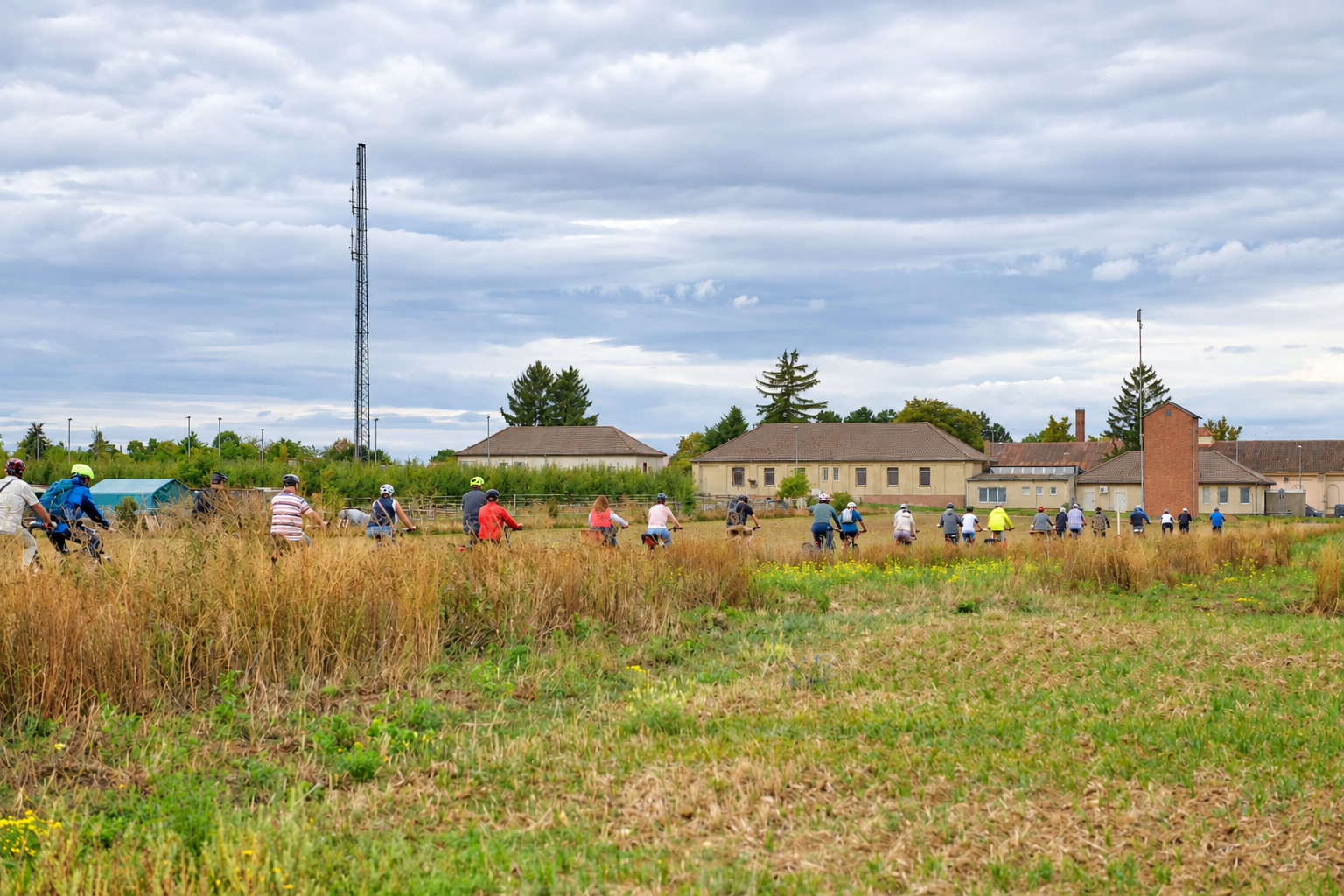 Zu sehen ist eine Aufnahme der Rheinhessischen Veranstaltung "Wasserradeln". Eine Gruppe von Radfahrerinnen und Radfahrer fahren eine Straße entlang, links und rechts ist Feld und Wiesen. Der Himmel ist leicht bedeckt, in der Ferne sind einige Gebäude.
