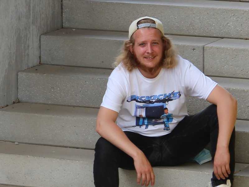 Marvin Mißkampf sitzt auf Betontreppen. Er hat lange, blonde Haare und trägt ein weißes T-Shirt mit einem grafischen Design darauf.