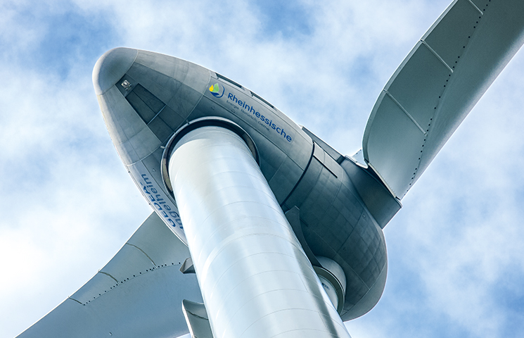 Nahaufnahme einer Windturbine vor einem blauen Himmel mit einigen Wolken. Der Fokus liegt auf der Gondel (das Gehäuse, das den Generator und andere Komponenten enthält) und dem Ansatz der Rotorblätter.