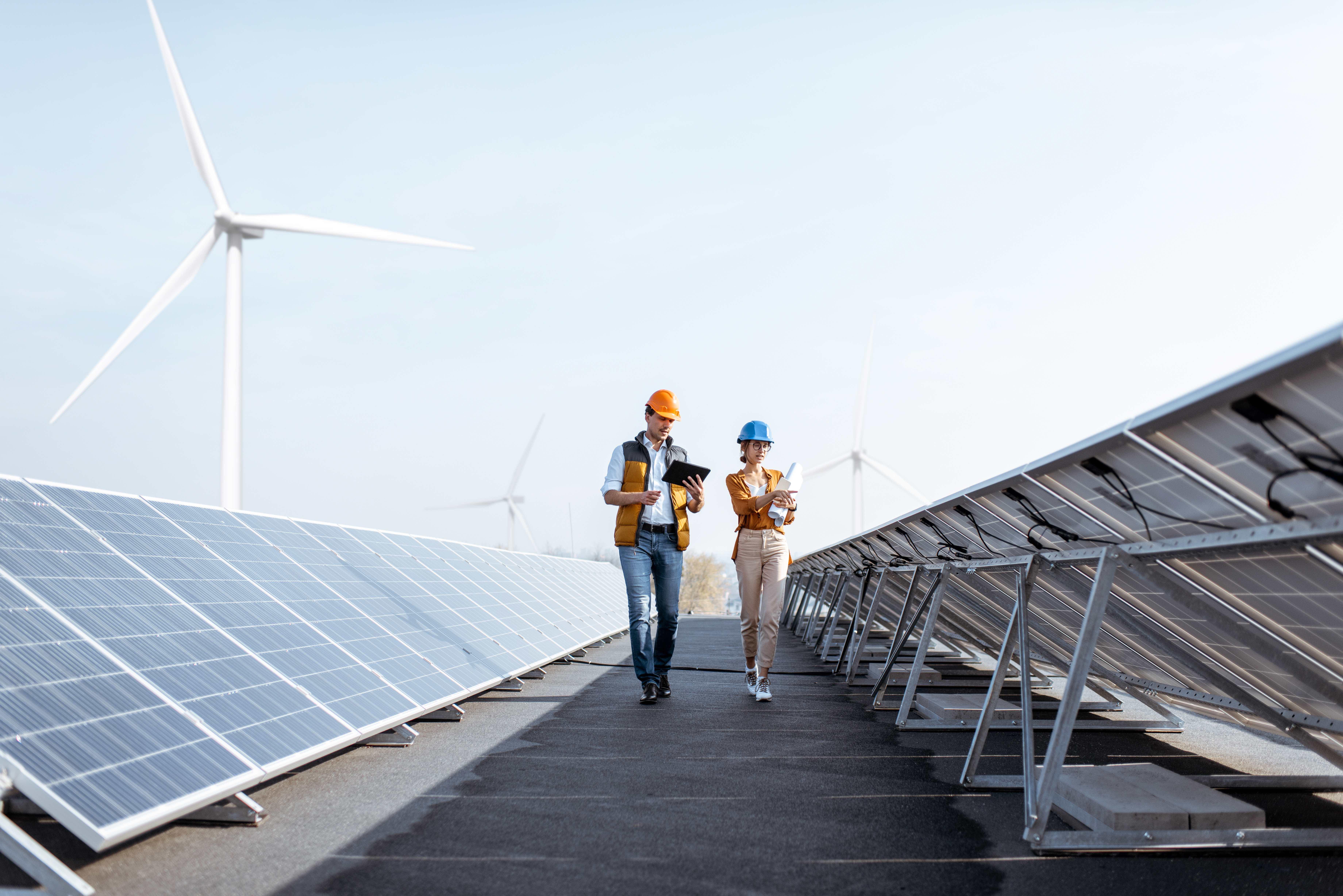 Das Bild zeigt zwei Personen, die Sicherheitshelme und Westen tragen und zwischen Reihen von Solarpaneelen auf einer Solarfarm gehen. Im Hintergrund sind Windturbinen sichtbar.