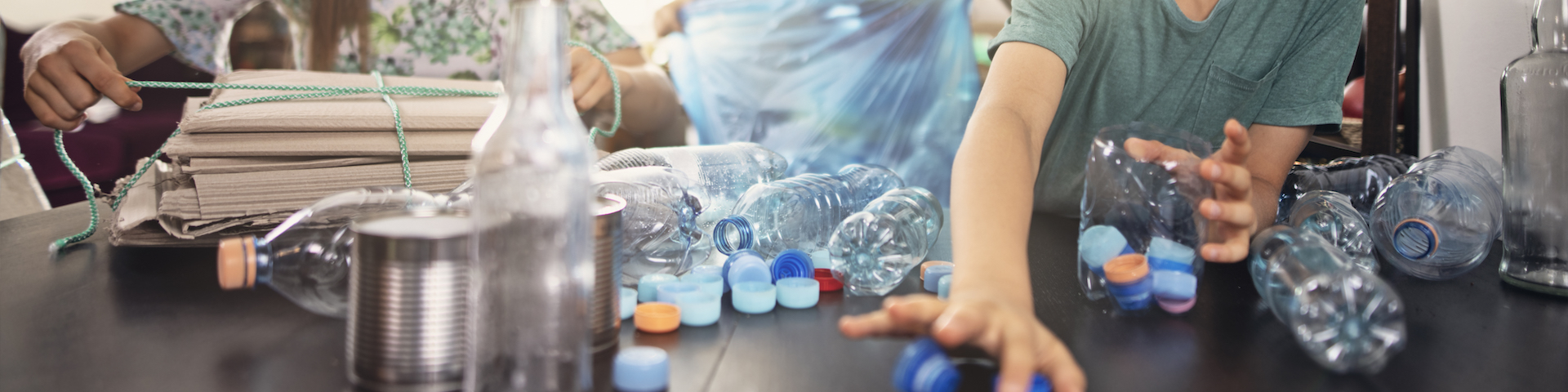 Das Bild zeigt eine Gruppe von Menschen, die verschiedene Plastikflaschen und -deckel auf einem Tisch sortieren. Auf dem Tisch liegen mehrere Plastikflaschen, Flaschendeckel und andere recycelbare Materialien verstreut.