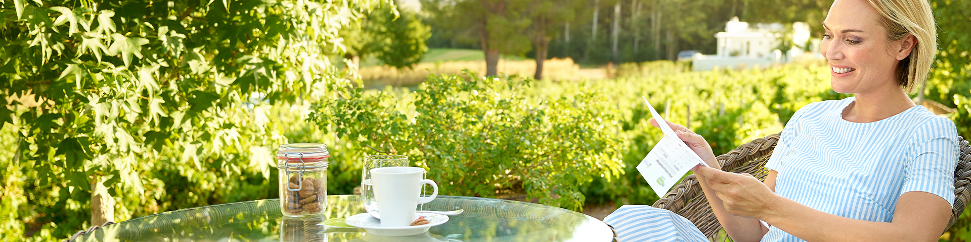 Das Bild zeigt eine Person, die draußen an einem Glastisch sitzt. Auf dem Tisch stehen eine Tasse Kaffee oder Tee und ein Glas mit Keksen. Die Person hält einige Papiere in der Hand und betrachtet sie.