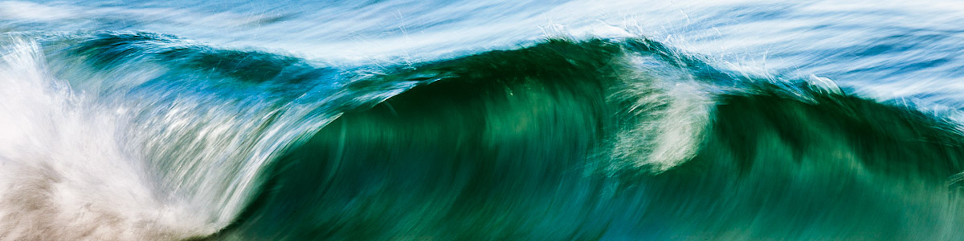 Das Bild zeigt eine große Ozeanwelle in verschiedenen Blau- und Grüntönen. Die Welle rollt sich über und erzeugt eine dynamische und kraftvolle Szene. Das Wasser scheint in Bewegung zu sein, und am Kamm der Welle bildet sich weißer Schaum.
