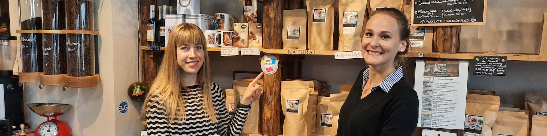Das Bild zeigt zwei Personen, die vor Regalen mit verschiedenen verpackten Produkten stehen. Eine Person zeigt auf einen runden Aufkleber am Regal.