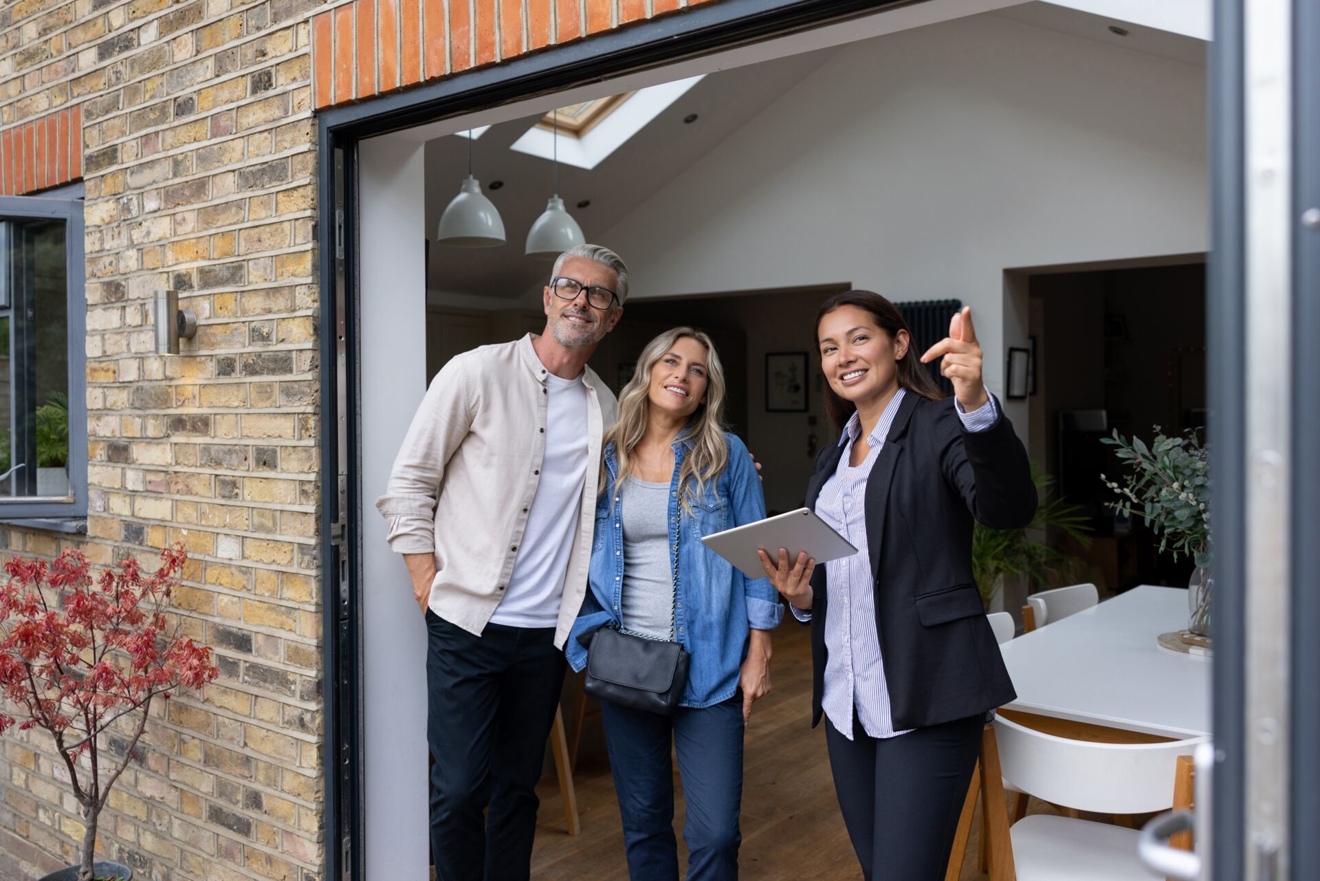 Drei Personen stehen am Eingang eines Hauses. Eine Person zeigt auf etwas im Haus, die anderen schauen zu. Die Umgebung ist modern mit Pendelleuchten, Holzböden und Möbeln. Es gibt auch etwas Grün im Haus und eine Ziegelwand außen.